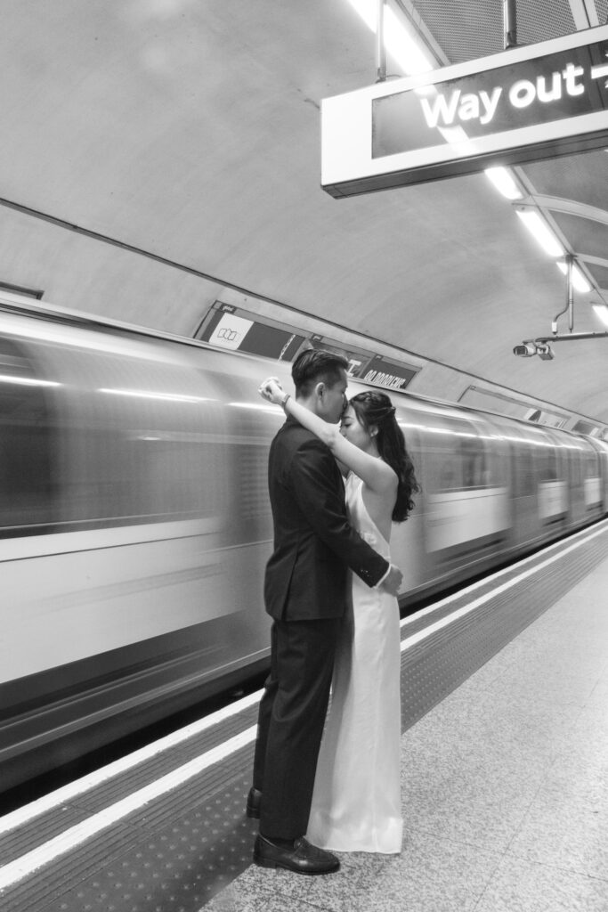 A couple dressed in formal attire embraces and kisses on a subway platform as a train speeds by in the background; a sign above reads Way out. The photo is in black and white.