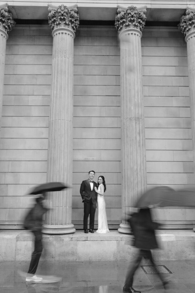 A couple in formal attire stands between tall columns, posing together. Blurred figures with umbrellas walk past in the foreground, suggesting rain. The image is black and white.