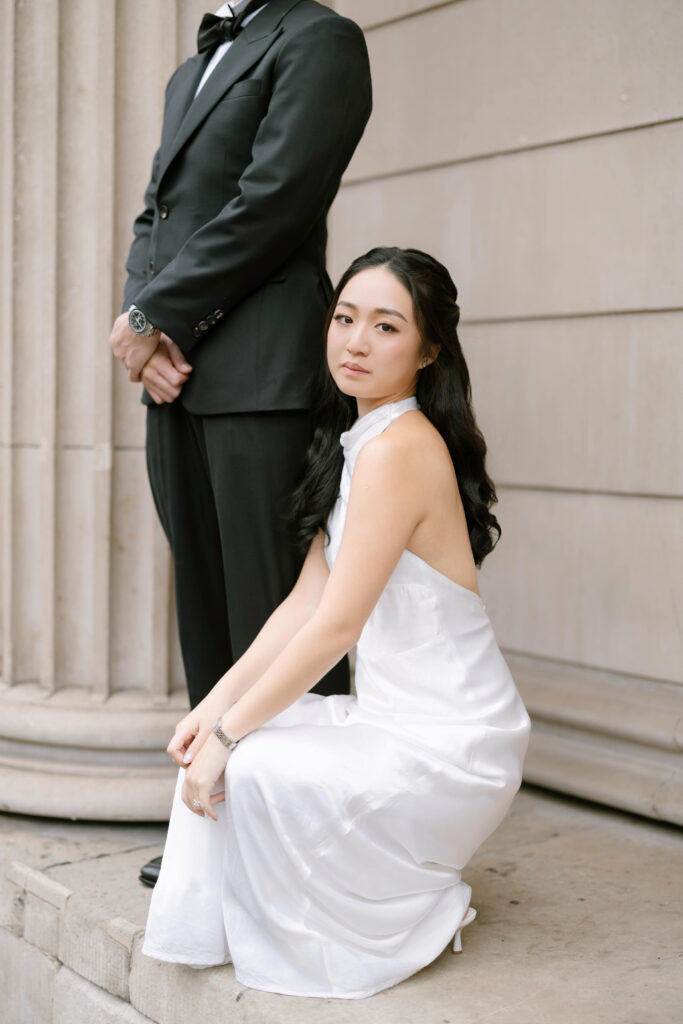 A woman in a sleeveless white dress sits on stone steps, looking at the camera, while a man in a black suit stands behind her with his hands clasped. They are in front of a building with large columns.