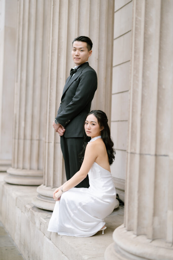 A woman in a white dress kneels beside a man in a dark suit standing, both posing against tall stone columns on a building exterior.