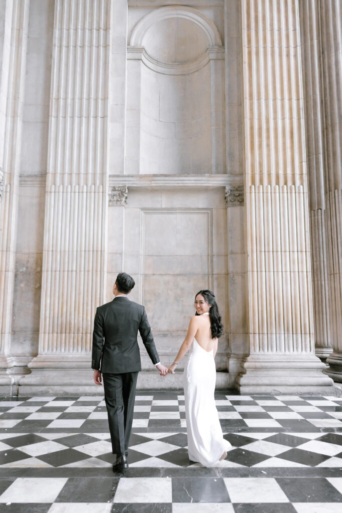 A couple holding hands walks away on a black and white checkered floor, surrounded by tall, ornate columns and large stone walls. The woman wears a white dress and looks back, while the man wears a dark suit.
