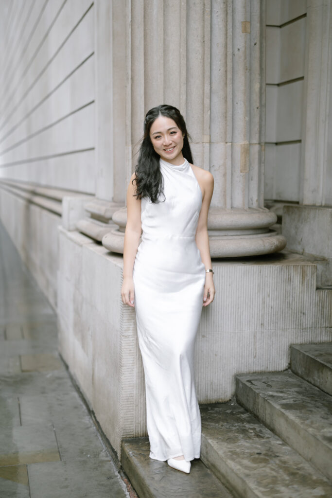 A woman with long dark hair in a white satin dress stands smiling by stone steps and large columns on a city sidewalk.