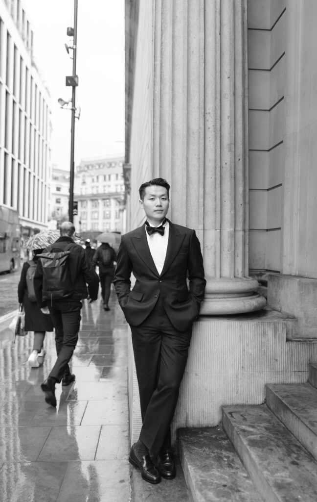 A man in a tuxedo leans against a large column on a city street, hands in pockets, looking away. Pedestrians walk by on the wet pavement under umbrellas. The scene is in black and white.