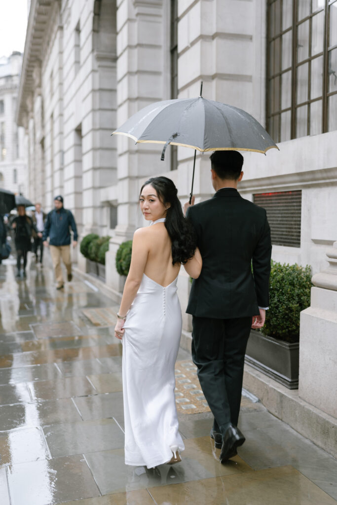 A woman in a white dress and a man in a black suit walk together on a rainy city street. The man holds a black umbrella over them. The woman looks back over her shoulder, and the pavement is wet from rain.