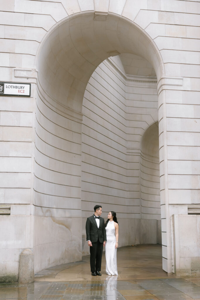 A couple dressed in formal attire stand beneath a large stone archway, looking at each other, with the street sign Lothbury EC2 visible on a building in the background. The ground appears wet, suggesting recent rain.