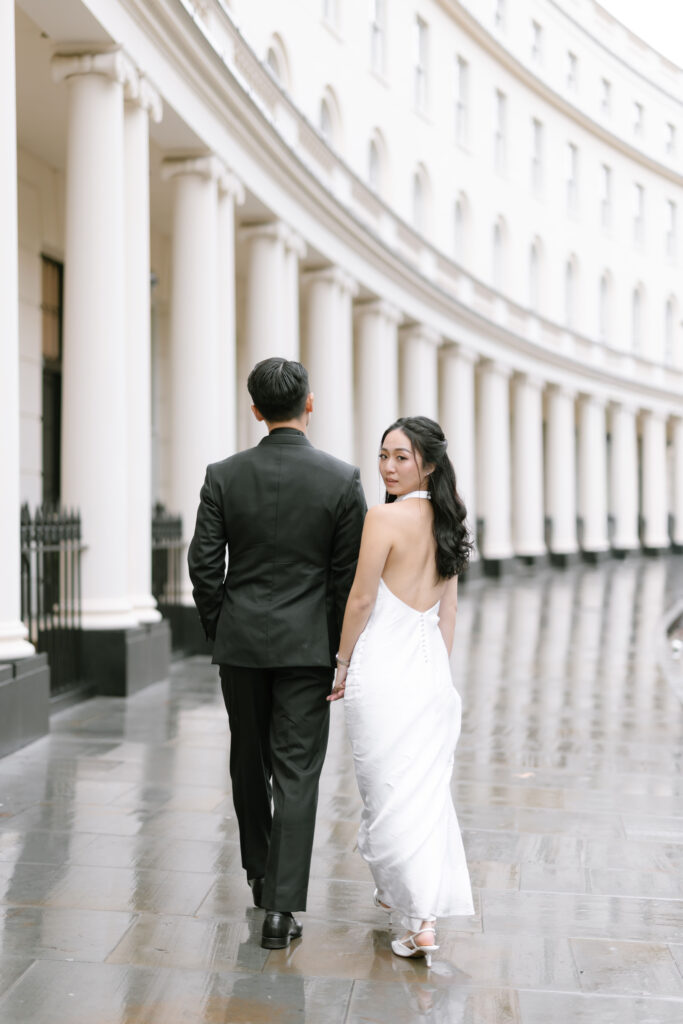 A couple dressed in formal attire, with the woman in a white backless gown and the man in a black suit, walk hand-in-hand along a curved, column-lined building on a wet pavement. The woman looks back over her shoulder.