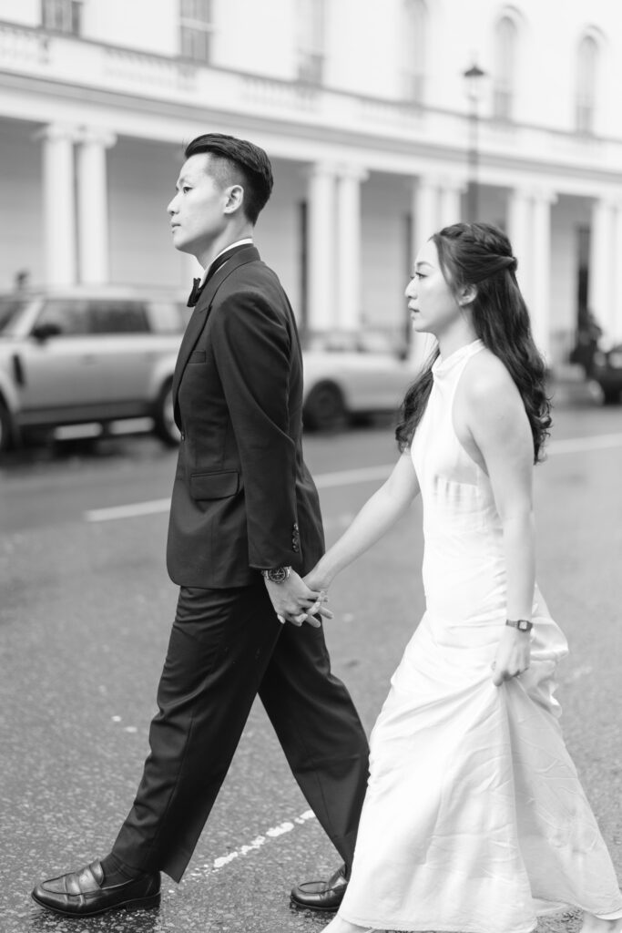 A couple dressed formally, with the man in a suit and the woman in a white dress, walk hand in hand across a city street on a rainy day. The background shows parked cars and a building with columns.