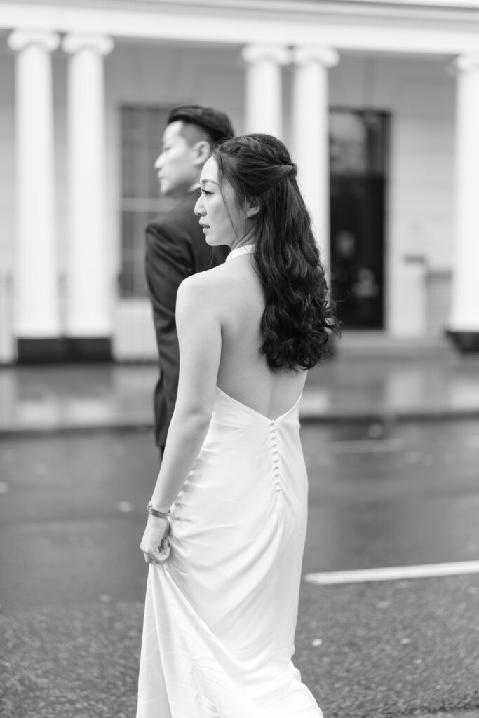 A woman in a backless white dress walks alongside a man in a suit on a city street, with white columns and blurred buildings in the background. The image is in black and white.