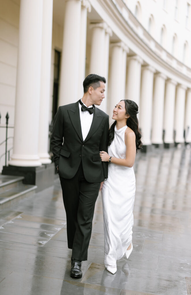 A couple dressed formally, the man in a black tuxedo and bow tie and the woman in a white gown, walk arm in arm on a wet sidewalk in front of a curved building with tall columns, smiling at each other.