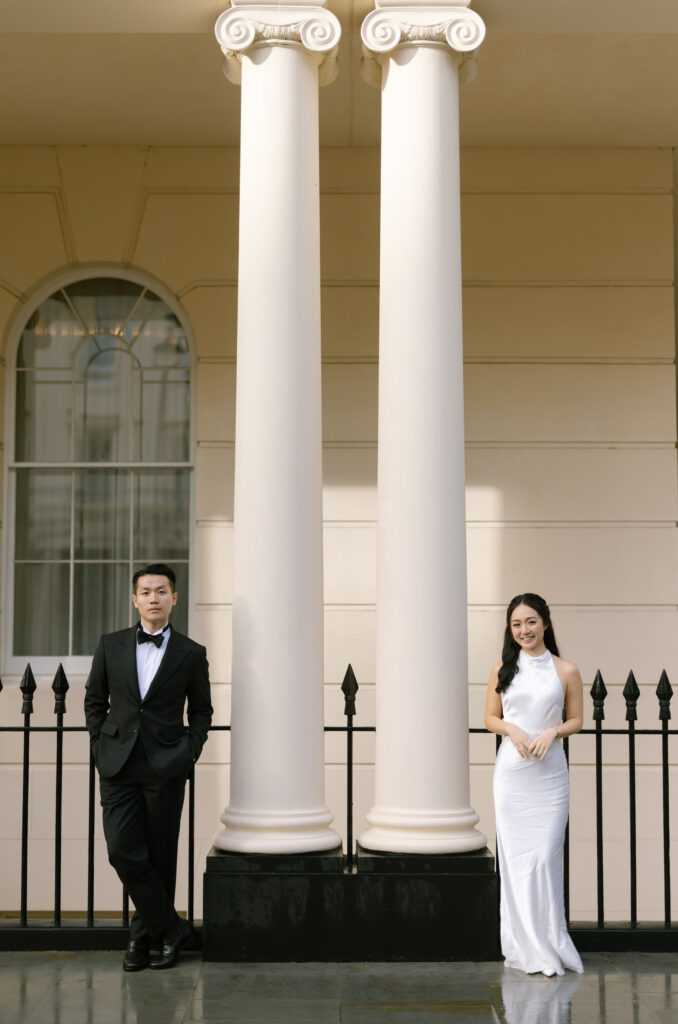 A man in a black suit and a woman in a white dress stand on either side of two large white columns in front of a building with arched windows and a black iron fence.
