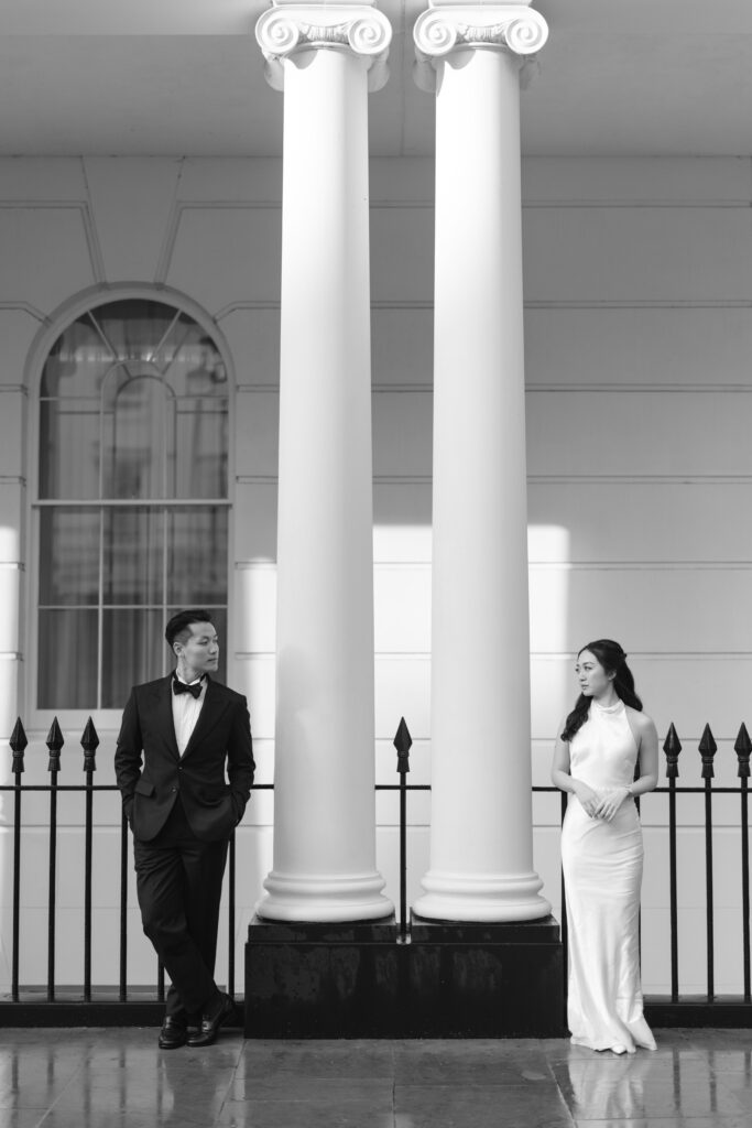 A man in a tuxedo and a woman in a white dress stand on either side of two large columns, facing each other, in front of a building with a tall arched window and black fence. The image is in black and white.