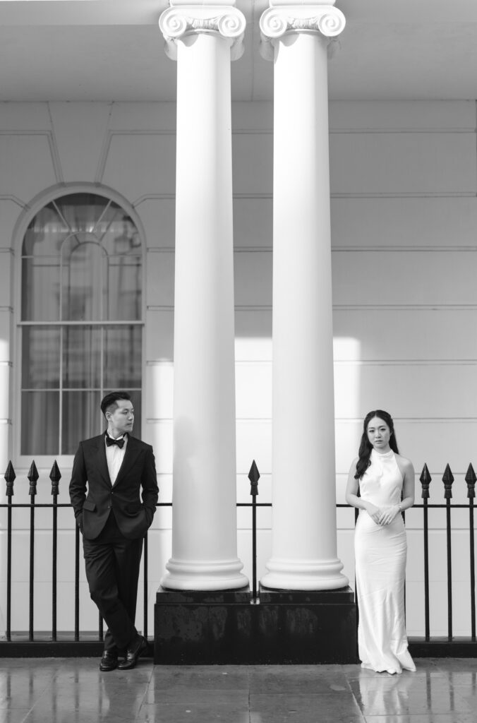 A man in a tuxedo and a woman in a white dress stand apart on either side of two large white columns outside a classical building, each looking in opposite directions. The scene is elegant and monochrome.