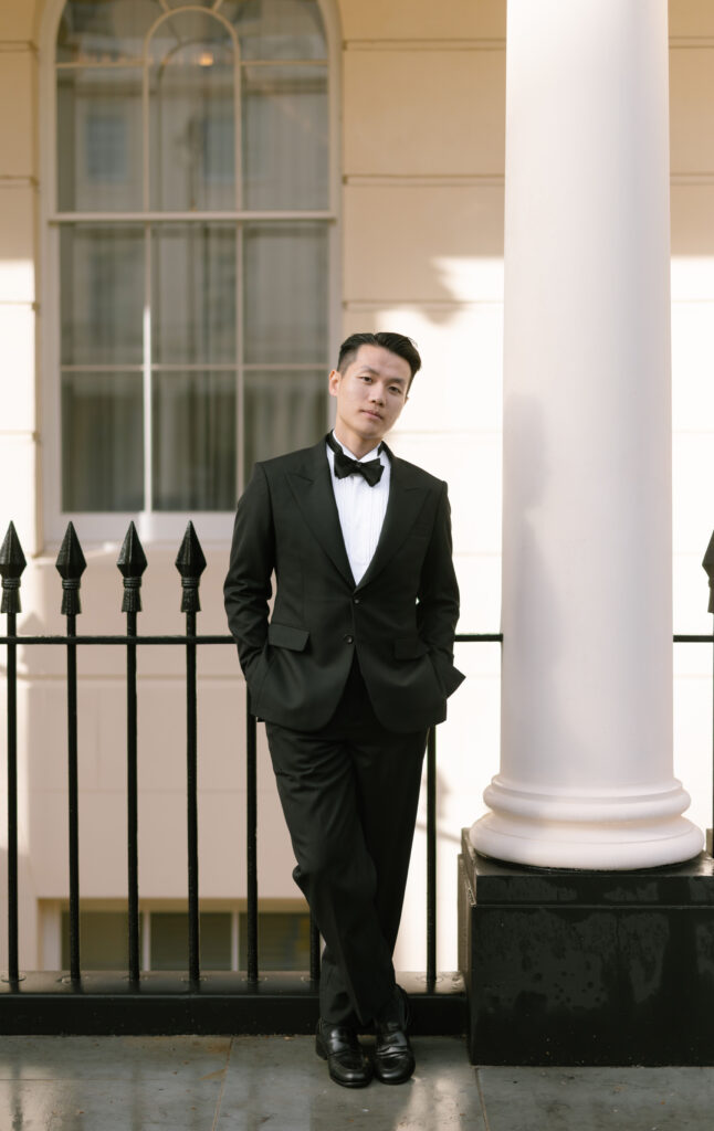 A man in a black tuxedo and bow tie stands with hands in pockets, leaning slightly against a white column outside a building with large windows and black iron railings.
