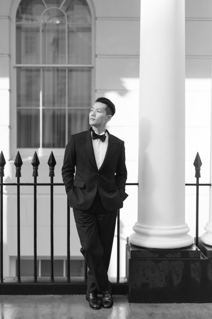 A man in a suit with a bow tie stands next to a large column, hands in pockets, and looks up while smiling. The background features a window, railings, and bright sunlight. The image is in black and white.