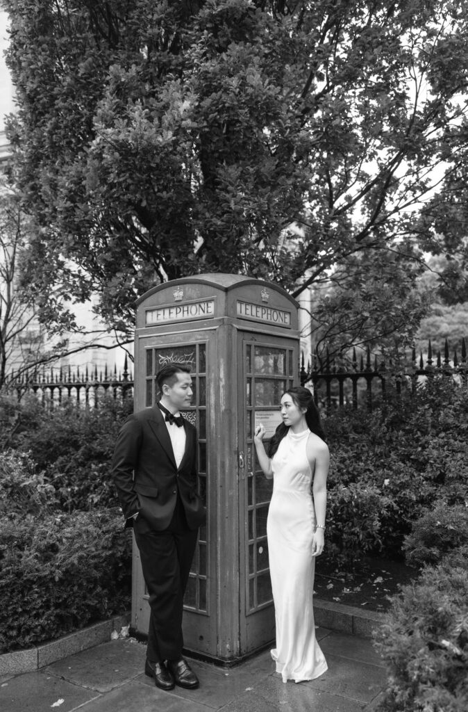 A man in a tuxedo and a woman in a white dress stand on either side of a classic British red telephone booth, surrounded by trees and greenery, in a black and white photo.
