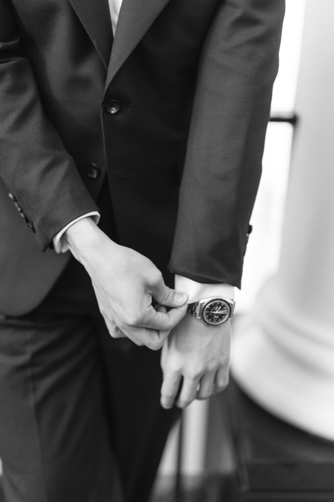 Black and white photo of a person in a suit adjusting their cuff with one hand, wearing a wristwatch. Their face is not visible, focusing on their torso and hands.