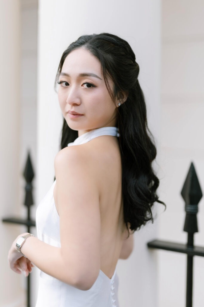 A woman with long, wavy black hair in a sleeveless white dress looks over her shoulder at the camera, standing outdoors in front of a white pillar and black metal fence.