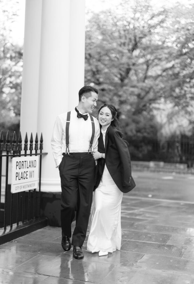 A couple dressed in formal attire walks together, smiling and laughing, on a wet sidewalk near a sign that reads Portland Place W1. The scene is outdoors with blurred trees in the background.