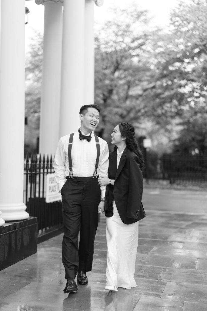 A couple walks arm in arm on a wet sidewalk, smiling at each other. The man wears dress pants, a shirt, suspenders, and a bow tie. The woman wears a long dress and a blazer. Tall columns and trees are in the background. Black and white photo.