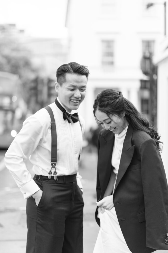 A black-and-white photo of a smiling couple standing outdoors. The man wears a bow tie, suspenders, and dress shirt, while the woman wears a blazer over a dress. Both are looking down, appearing cheerful and relaxed.