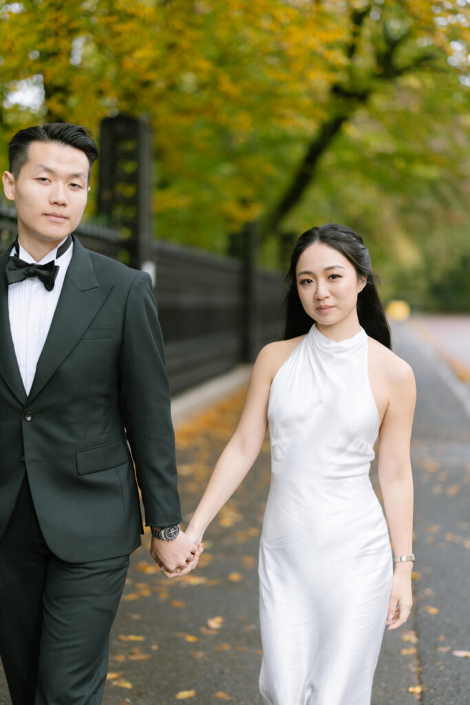 A man in a black tuxedo and a woman in a white sleeveless dress hold hands while walking on a tree-lined path with autumn leaves.