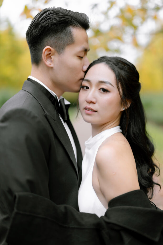 A man in a black suit kisses the forehead of a woman in a white dress. They embrace outdoors, framed by greenery and soft, yellow autumn leaves in the background. The woman looks calmly at the camera.