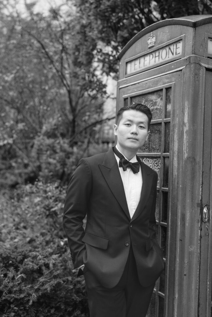 A person in a tuxedo stands with hands in pockets next to a vintage British red telephone booth, surrounded by trees and foliage. The image is in black and white.