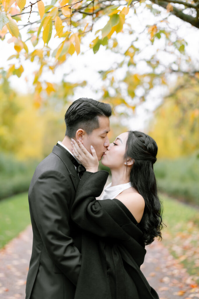 A couple dressed formally share a kiss under autumn trees with yellow leaves. The woman holds the mans face as they embrace on a pathway lined with greenery.