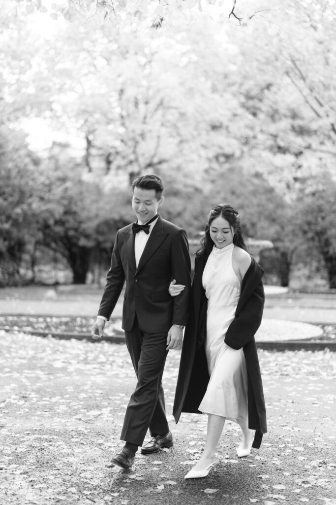 A couple dressed formally walks arm in arm outdoors on a tree-lined path. The man wears a suit and bow tie, while the woman wears a dress with a long coat. Fallen leaves cover the ground, giving an autumn feeling. Black and white photo.