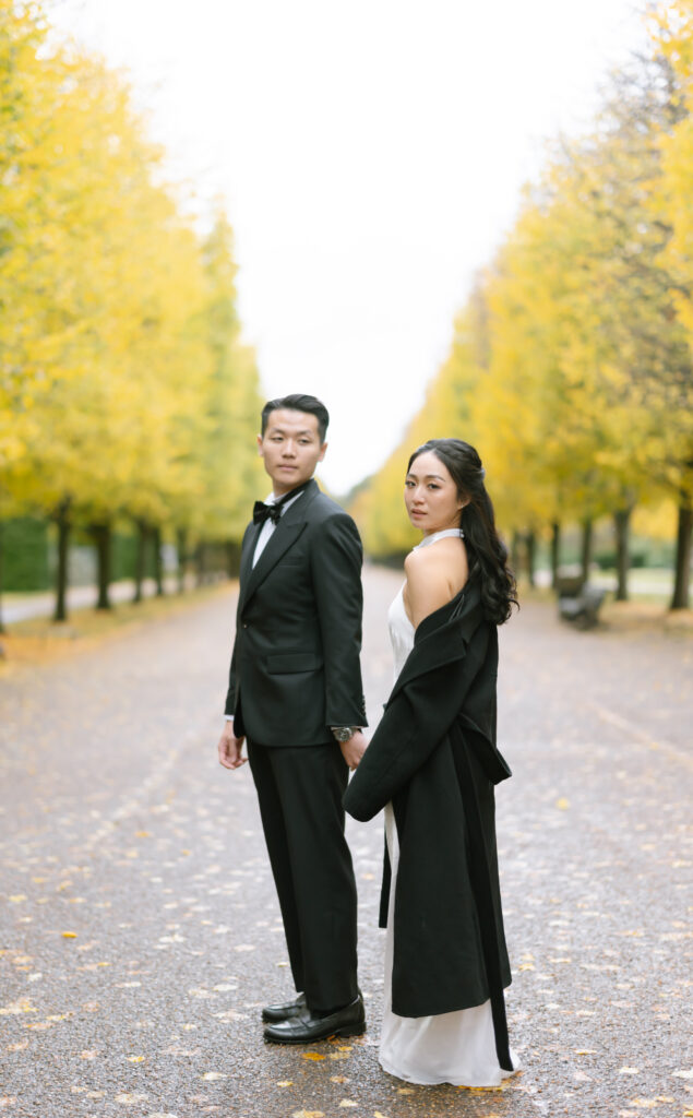 A couple dressed formally stands back-to-back on a tree-lined path with yellow autumn leaves. The man wears a black suit and bow tie; the woman wears a white dress and black coat. They look in opposite directions.