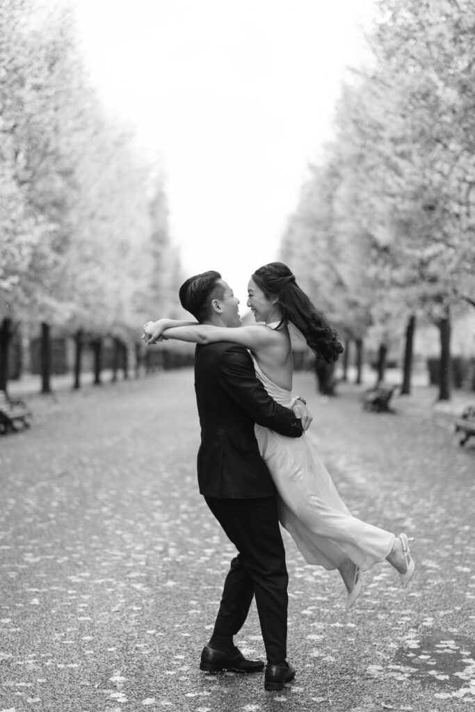 A man in a suit lifts a woman in a long dress as they smile at each other on a tree-lined path covered in fallen leaves. The scene is captured in black and white.
