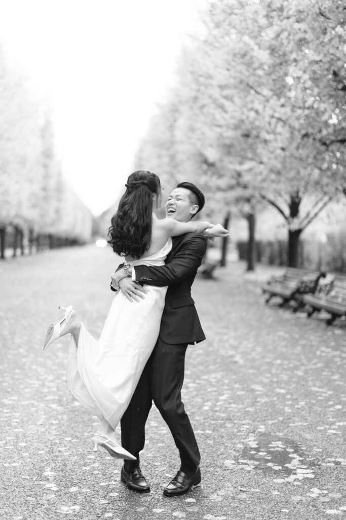 A joyful couple, dressed in formal attire, embrace on a tree-lined path. The man lifts the woman off the ground as they laugh together amid scattered petals. The scene is captured in black and white.