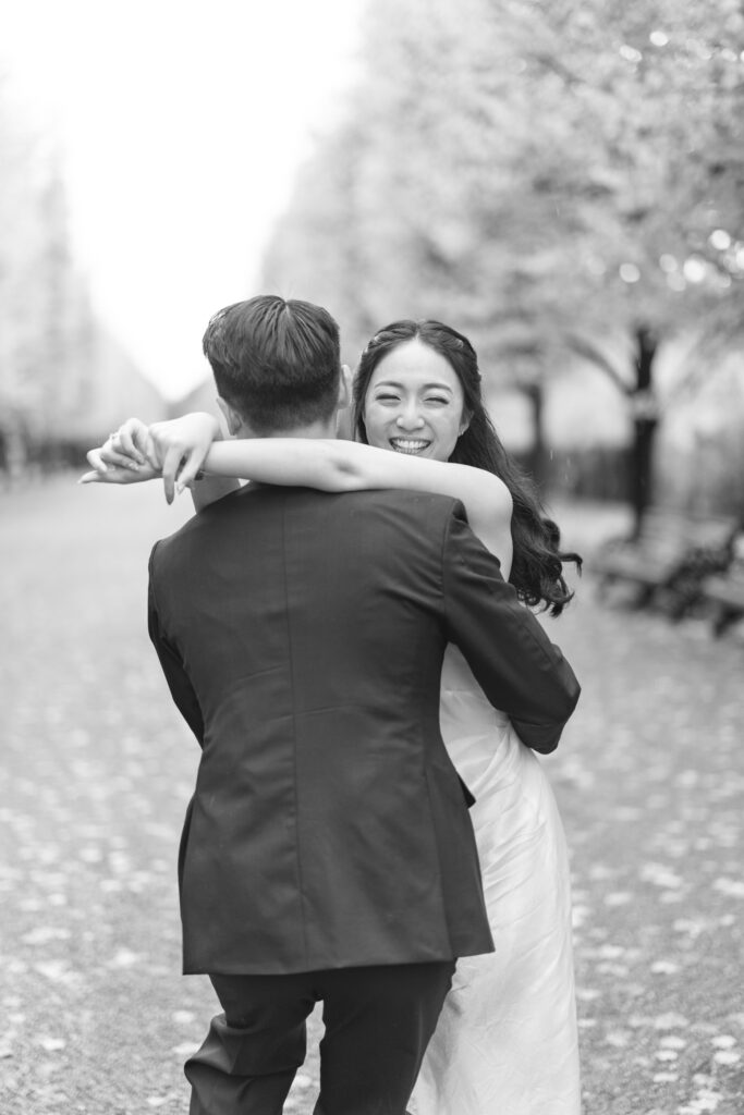 A joyful couple dances outdoors on a tree-lined path. The woman in a white dress smiles brightly, embracing the man in a suit, with autumn leaves scattered on the ground. The scene feels lighthearted and romantic.