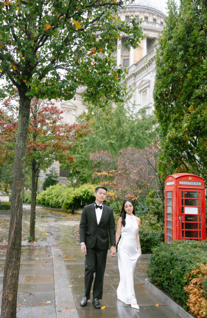 A couple dressed in formal attire walks hand-in-hand on a wet sidewalk near a red British phone booth and lush greenery, with a large historic building in the background.