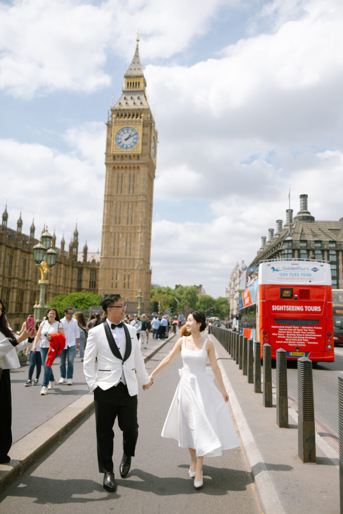 A couple in formal white and black attire walk hand-in-hand on a street in front of Big Ben in London, smiling and surrounded by tourists, with a red sightseeing tours sign visible nearby.