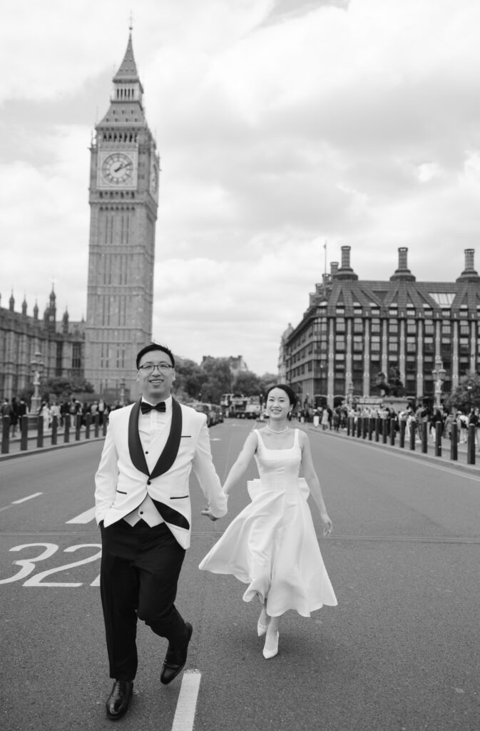 A bride and groom hold hands and run joyfully down a street in front of Big Ben in London. The groom wears a tuxedo, and the bride wears a white dress. The scene is lively and romantic, with historic buildings in the background.