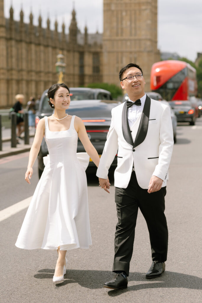 A couple dressed in formal wedding attire walks hand in hand on a street near the Palace of Westminster in London, with a red double-decker bus and cars in the background.