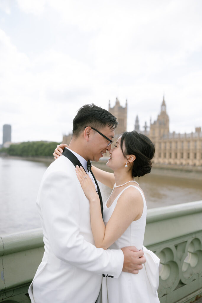 A couple in white formal attire embraces and smiles at each other on a bridge, with the Palace of Westminster and the River Thames in the background.