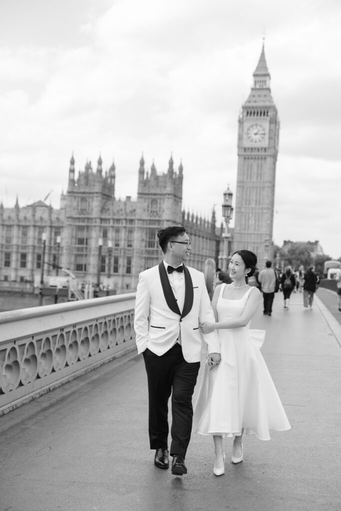 A couple dressed in formal wedding attire walks hand-in-hand on a bridge in front of Big Ben and the Houses of Parliament in London. The photo is black and white, with a joyful, romantic atmosphere.