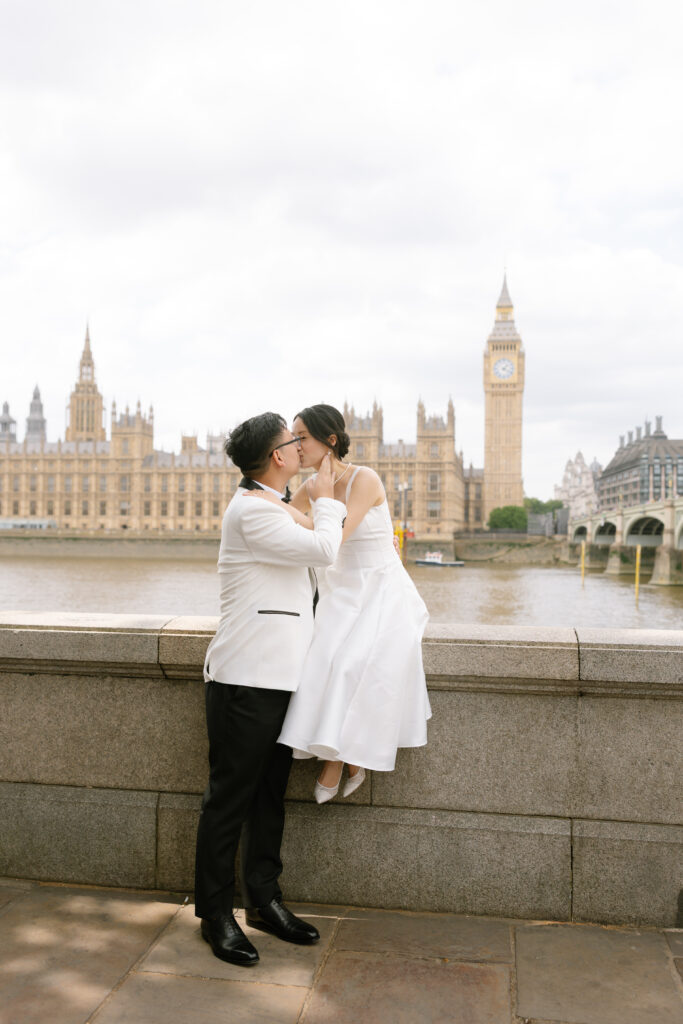 A couple dressed in formal wedding attire kisses while standing and sitting on a stone wall, with the River Thames and Big Ben in London visible in the background.