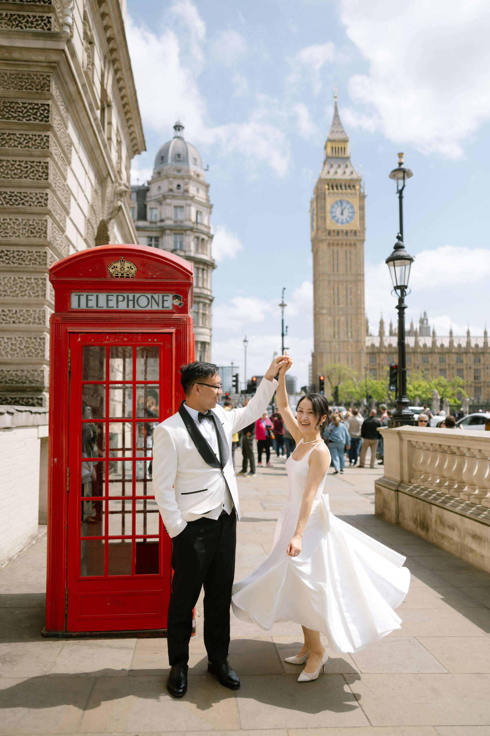 A couple in formal attire dance joyfully near a classic red telephone booth in London, with Big Ben and the Palace of Westminster in the background on a sunny day.