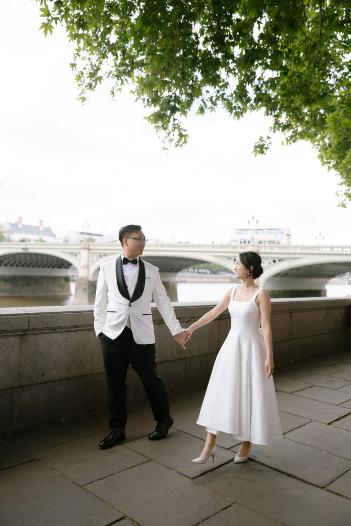 A couple dressed in formal wedding attire holds hands and smiles at each other while standing on a riverside walkway, with a large bridge and leafy tree overhead in the background.