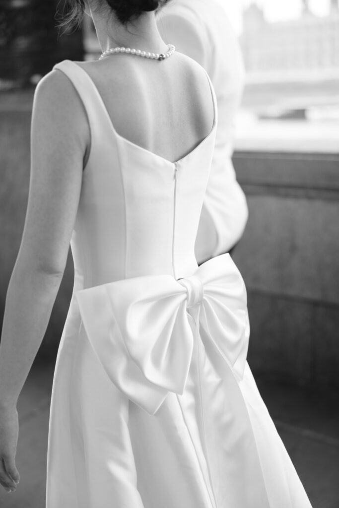 A woman in a sleeveless white dress with a large bow on the back and a pearl necklace stands next to a person in white, seen from behind, in a black and white outdoor setting.