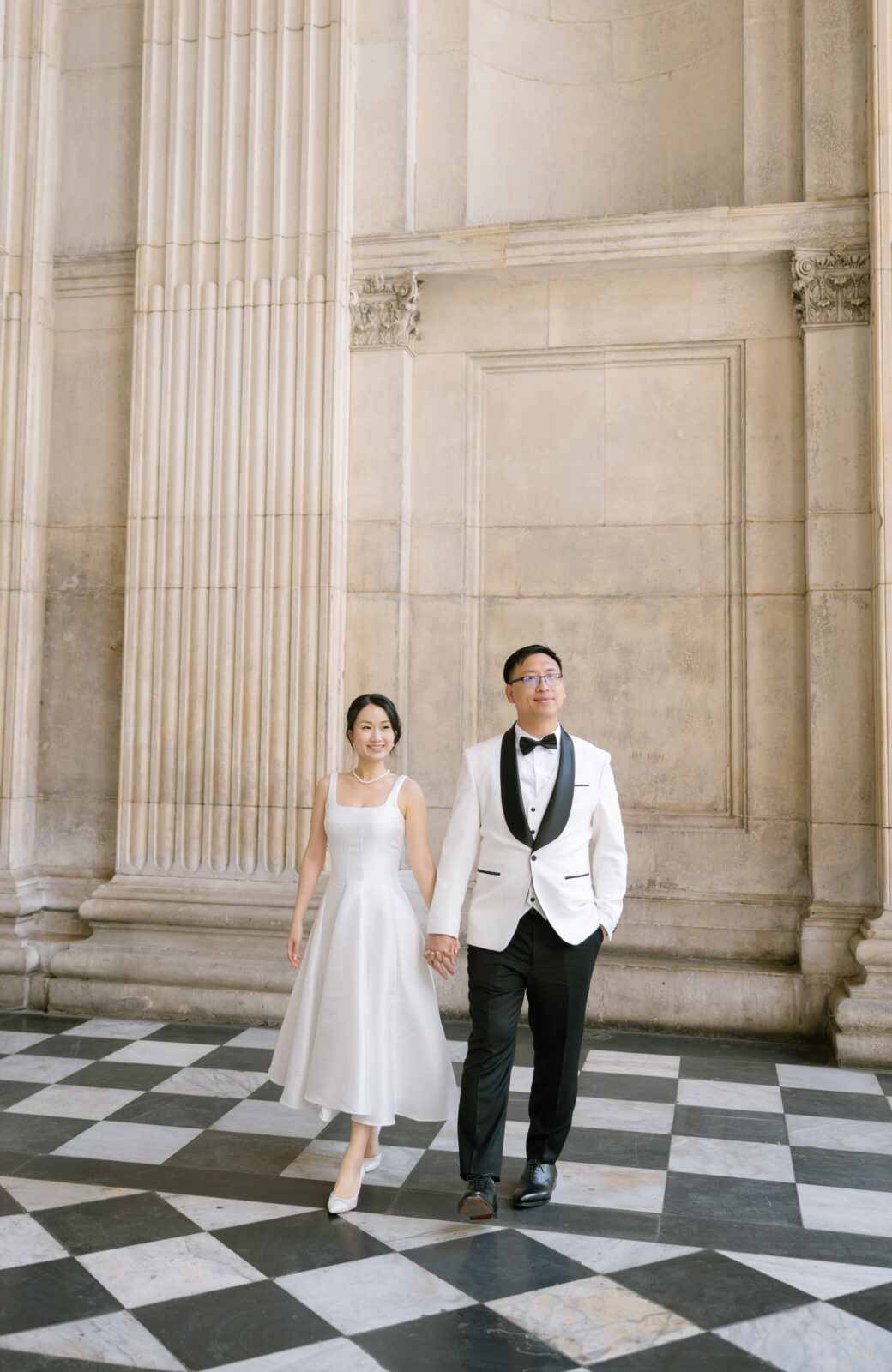 A couple dressed formally, with the woman in a white dress and the man in a white tuxedo jacket, walk hand in hand on a black-and-white checkered floor in front of tall, ornate columns.