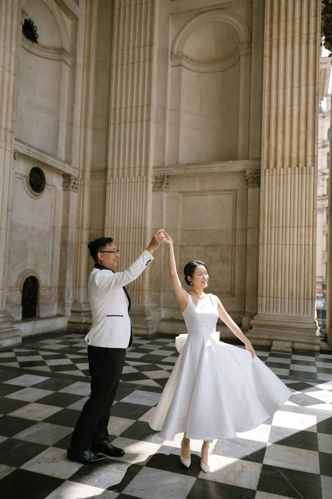 A couple dressed in formal attire dances on a black-and-white checkered floor in a grand, columned hall. The woman twirls in a white dress while the man, in a white jacket, holds her hand.
