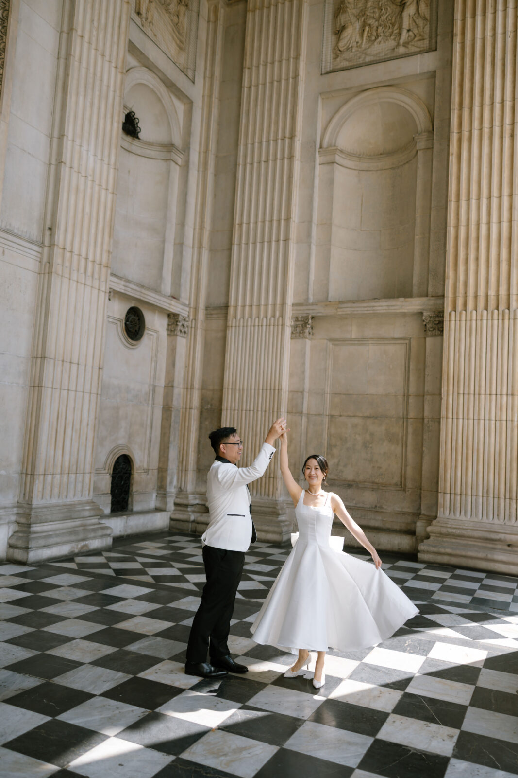 A couple dances together on a checkered black-and-white floor in an elegant, grand hall with tall columns and arched doorways. The woman wears a white dress, and the man is in a white jacket and black pants.