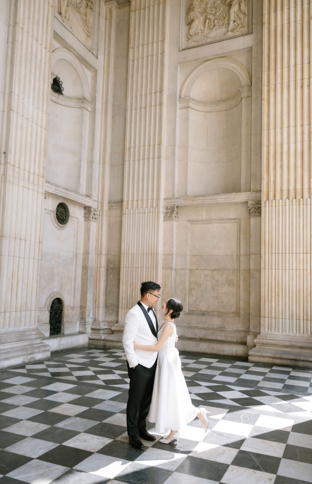 A couple dressed in wedding attire stands embraced on a black-and-white checkered floor in a grand, sunlit hall with tall columns and ornate architectural details.