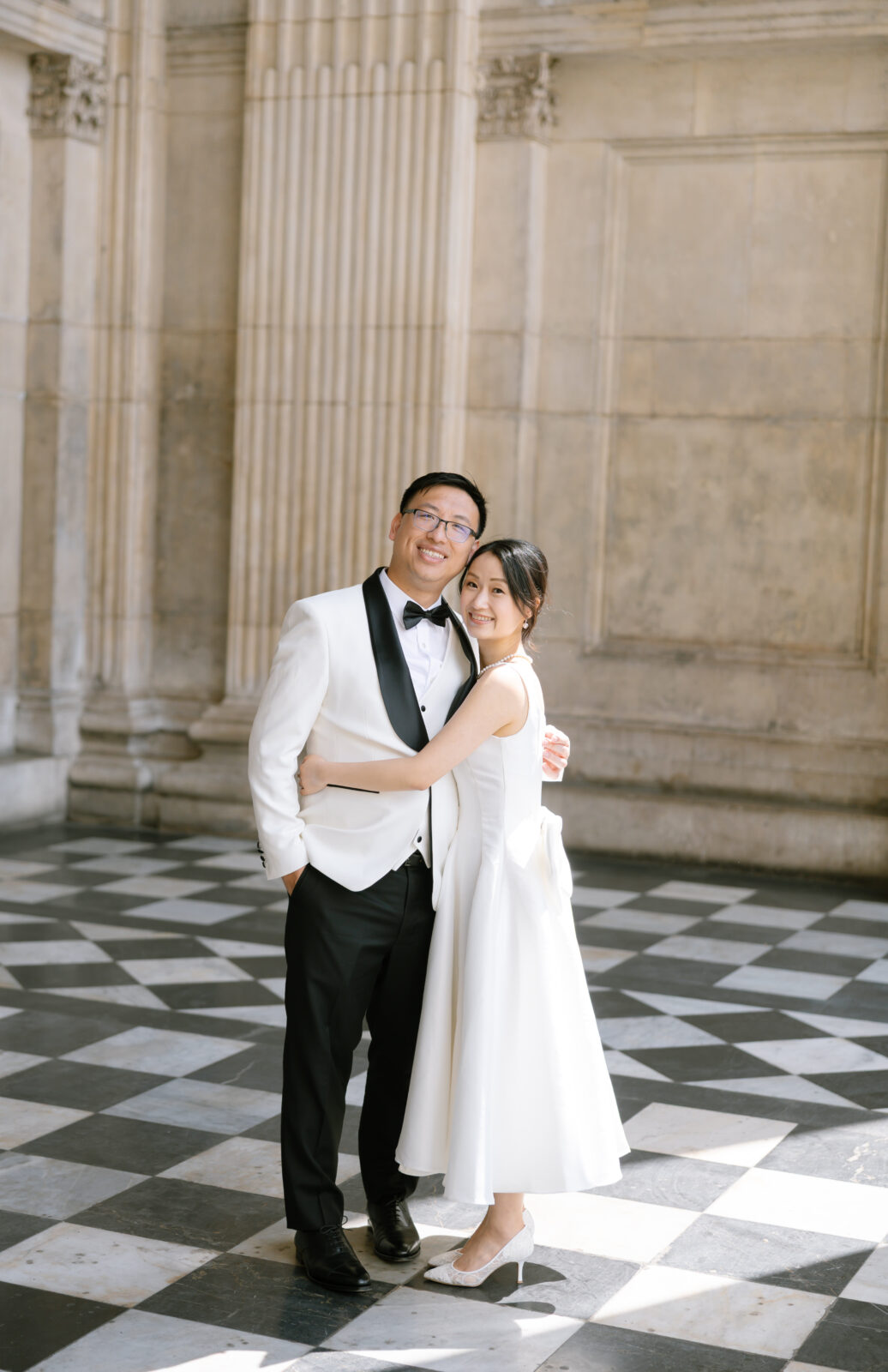 A couple dressed in formal attire, with the man in a white tuxedo and the woman in a white dress, stand smiling and embracing on a black-and-white checkered floor in front of tall, ornate columns.
