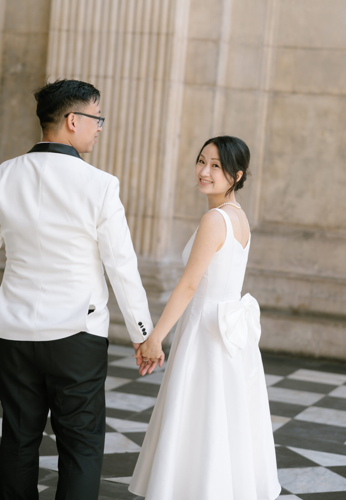 A couple in formal attire holds hands. The woman in a white dress with a bow looks back and smiles, while the man in a white jacket stands beside her. They are in front of a large stone wall on a checkered floor.