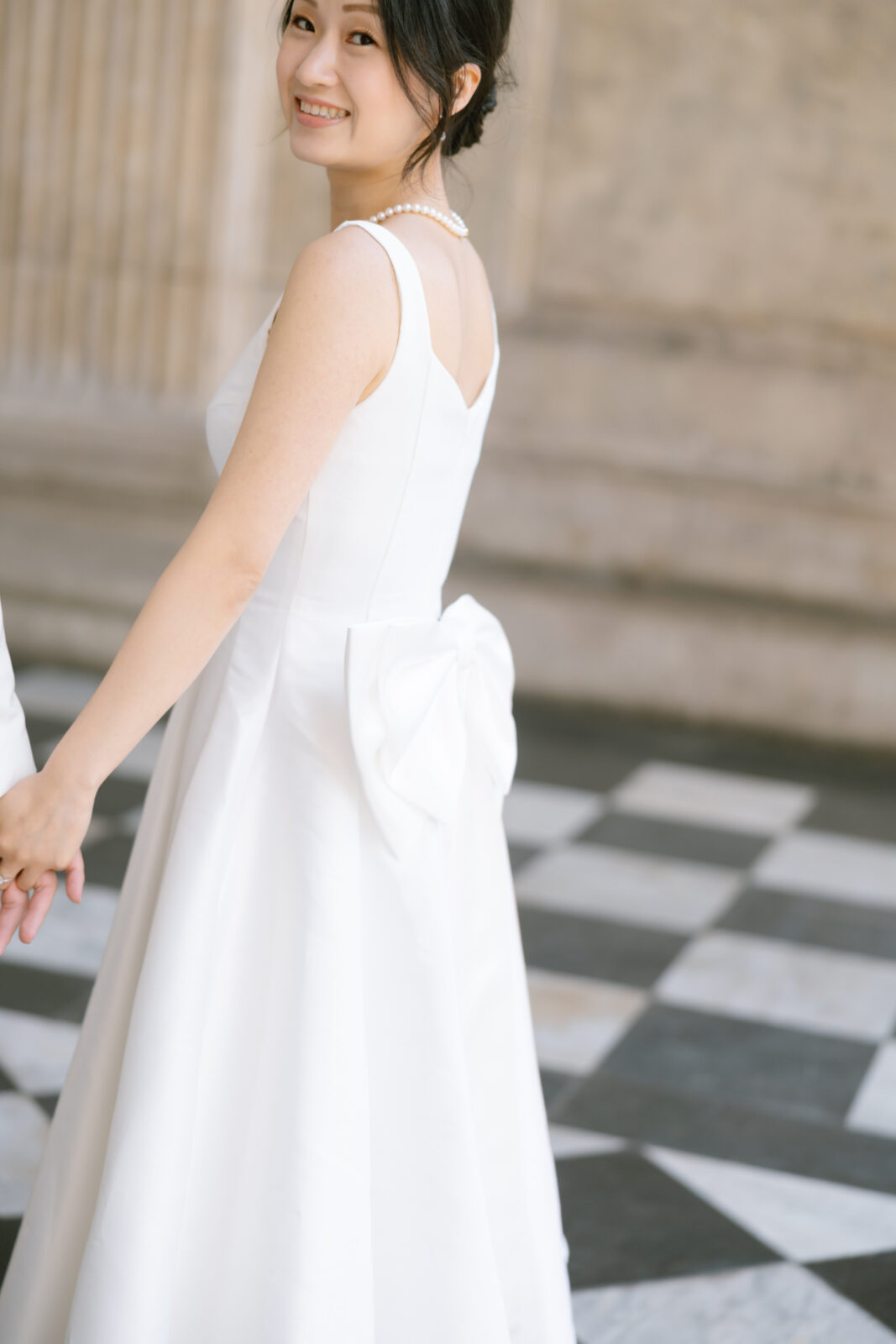 A smiling woman in a white sleeveless dress with a bow on the back holds hands with someone off-frame, standing on a black-and-white checkered floor in front of a stone wall.